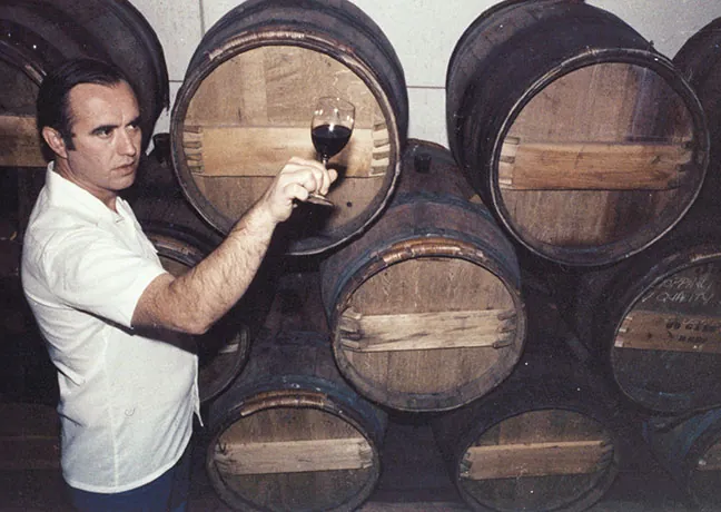 A man holding a glass of wine in front of barrels.