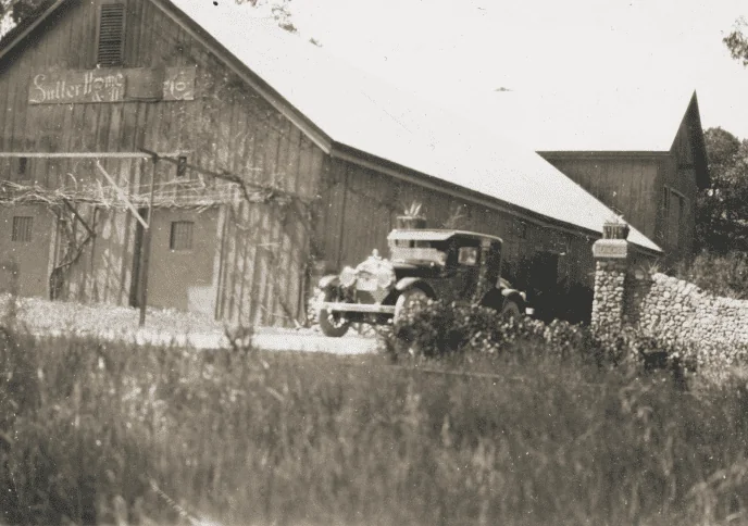A black and white photo of a barn with a car parked in front of it.