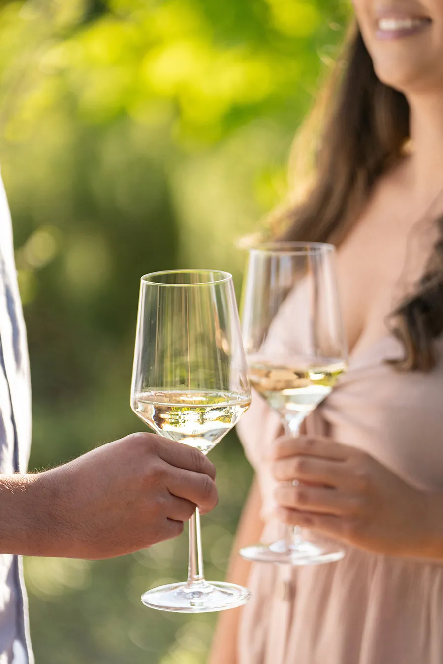 Two people holding glasses of white wine outside, with blurred greenery in the background.