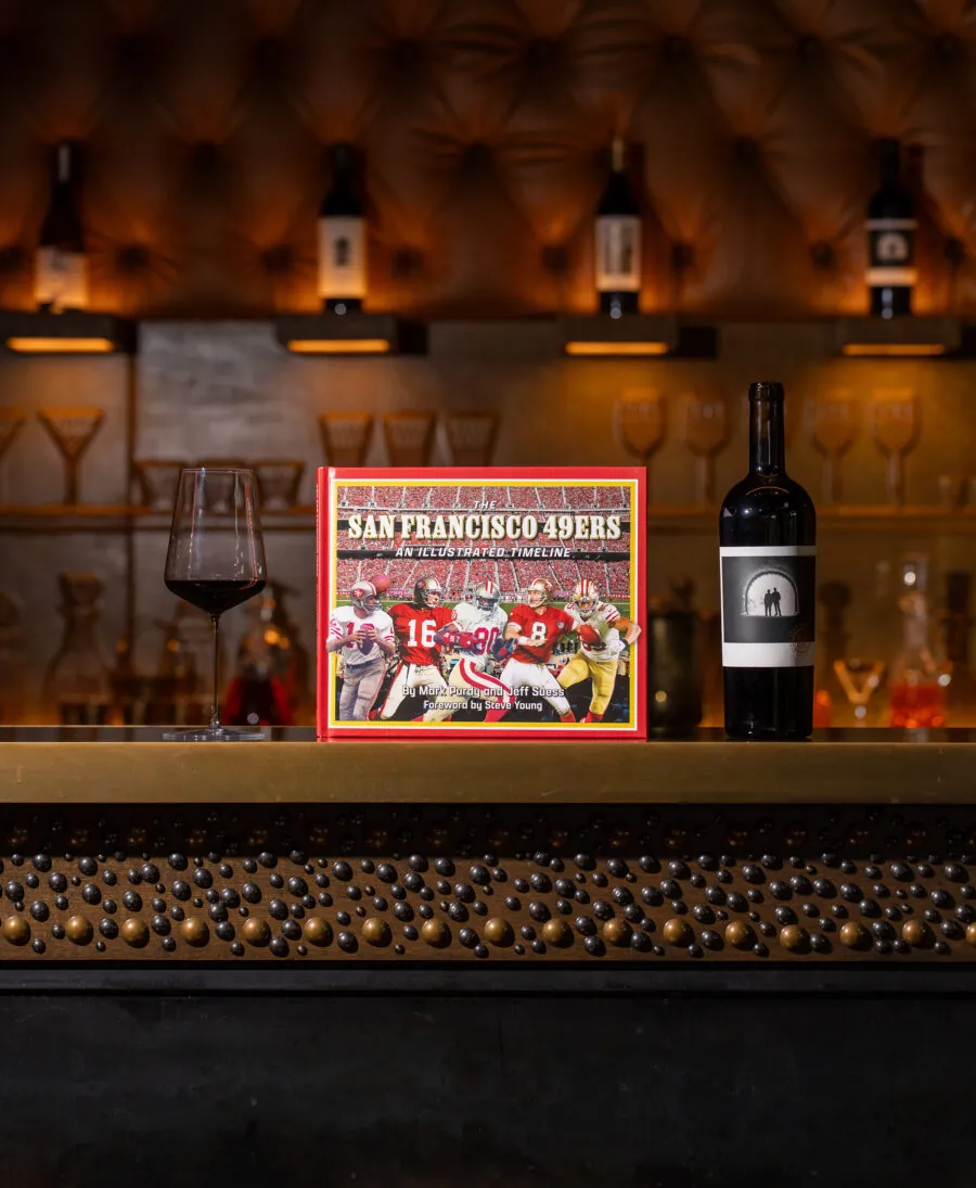 A San Francisco 49ers-themed book is displayed on a bar counter with a wine glass and a wine bottle; shelves with bottles and glasses are visible in the background.