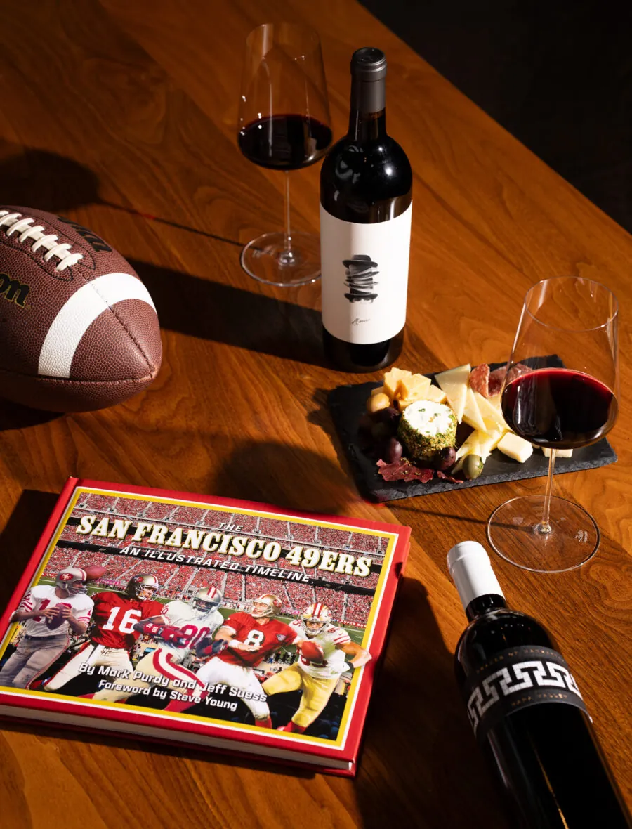 A San Francisco 49ers book, a football, two glasses and two bottles of red wine, and a slate plate with cheese and charcuterie are arranged on a wooden table.