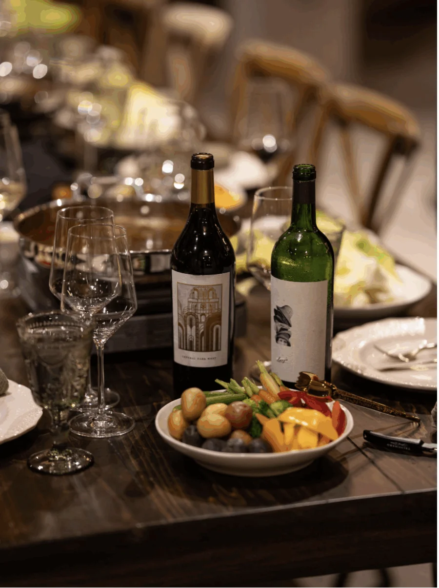 A wooden dining table set with wine bottles, empty wine glasses, a bowl of assorted vegetables, and plates, with other dishes and chairs in the background.