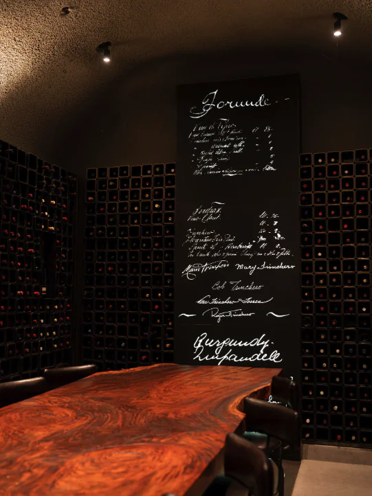 A wine cellar with a wooden table, black chairs, a wall of wine bottles, and a large blackboard listing wine options in white script.