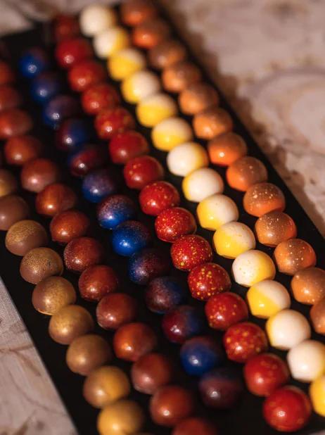 Rows of round, glossy, multicolored chocolates are neatly arranged on a black tray, featuring shades of blue, red, yellow, brown, orange, and white.