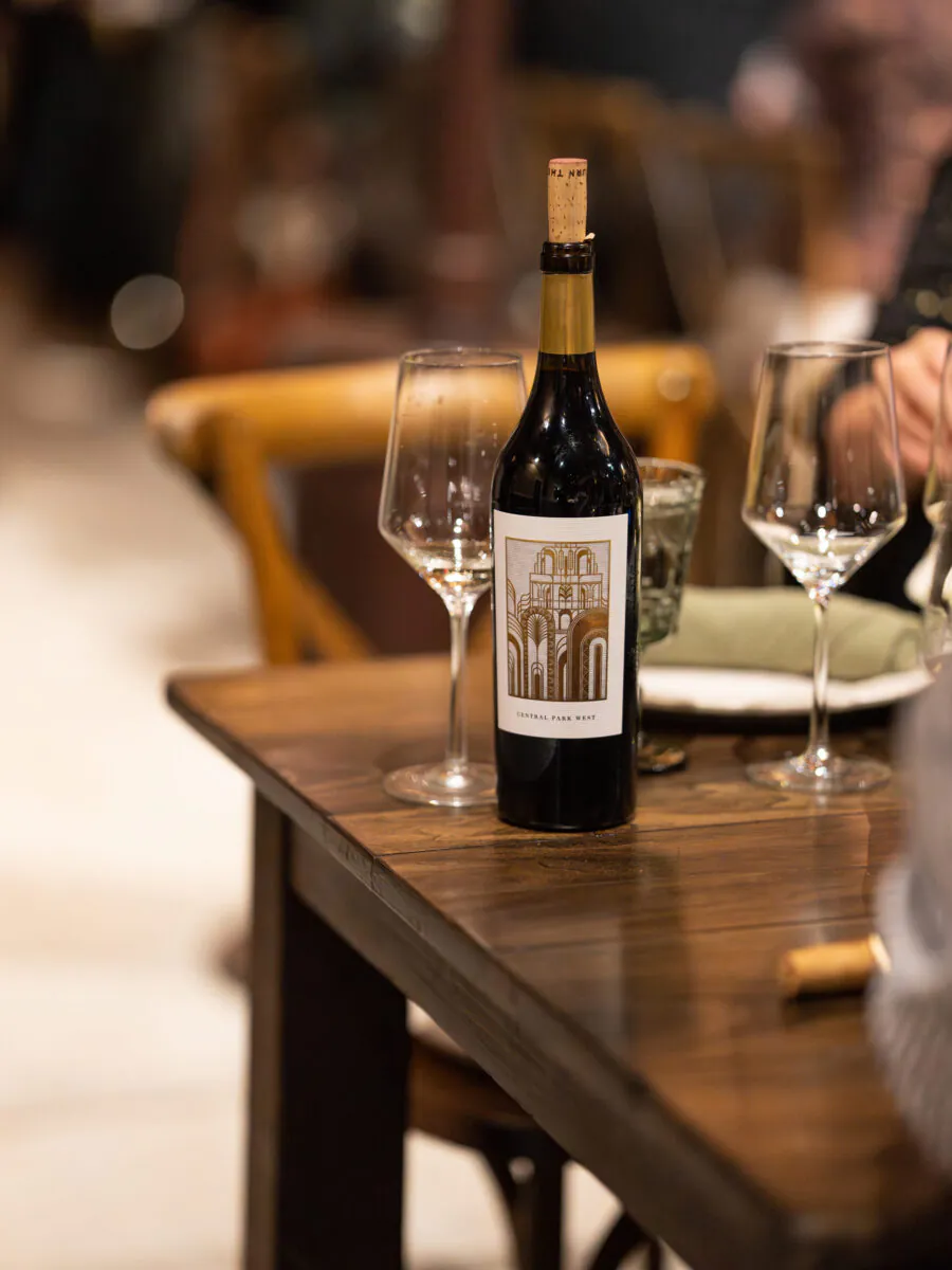 A bottle of red wine with a cork, two empty wine glasses, and a glass tumbler are on a wooden table in a restaurant setting.