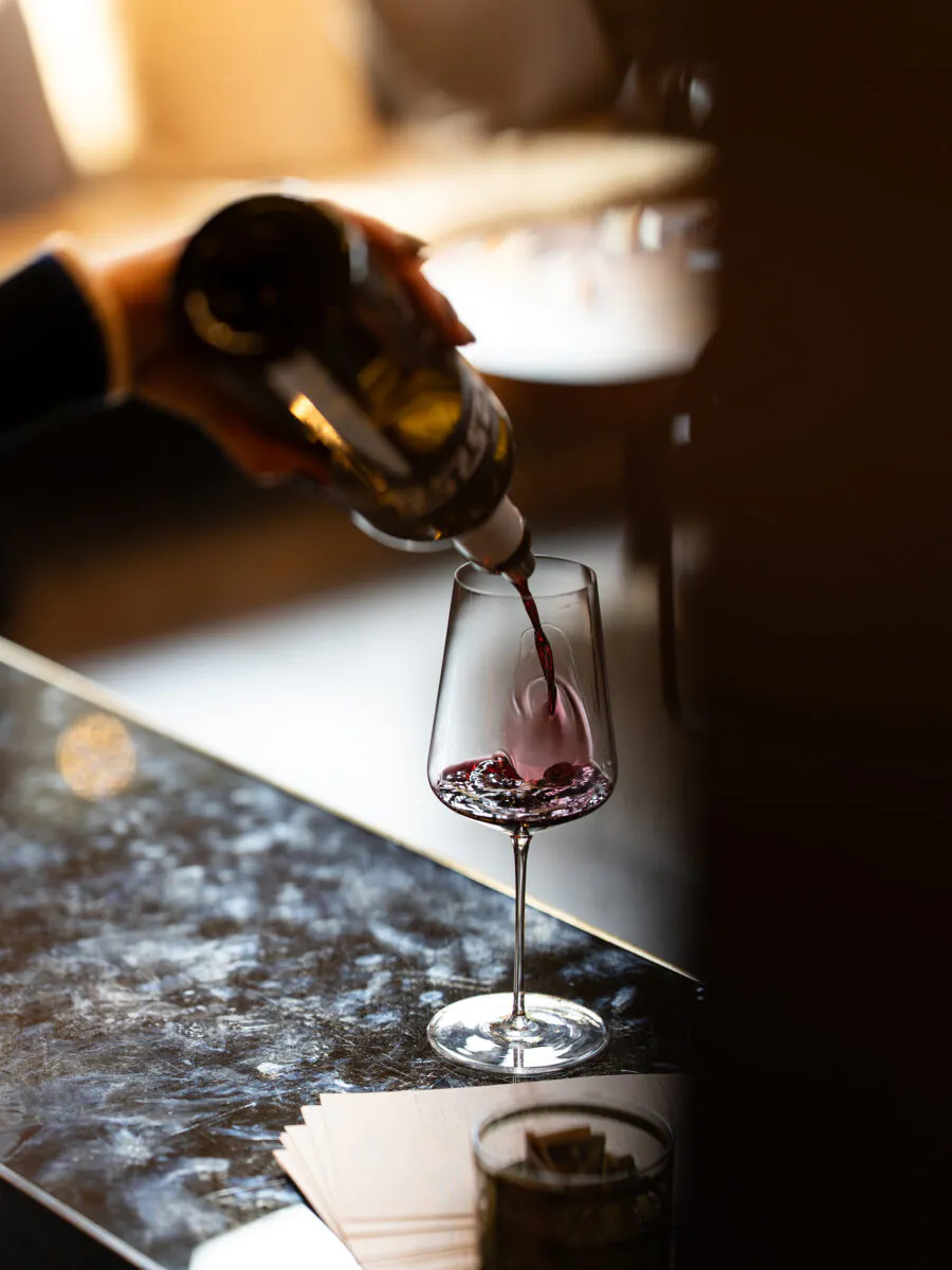 A hand pours red wine from a bottle into a wine glass on a marble countertop, with papers and a small container nearby.