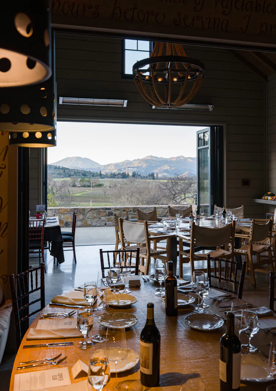 A restaurant dining area with set tables, wine bottles, and glassware overlooks a scenic vineyard and mountains through open doors.