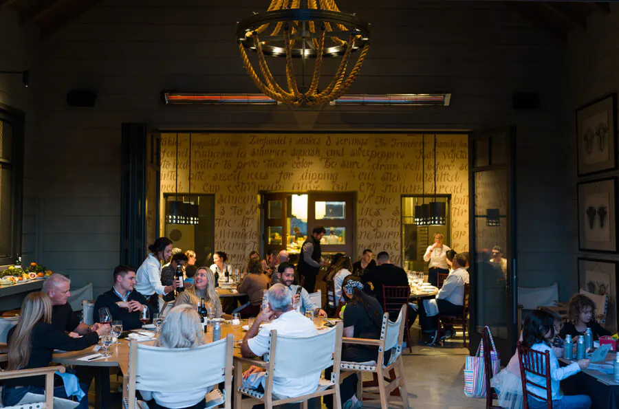 People dining at tables inside a warmly lit restaurant, with a chandelier above and writing on the back wall.
