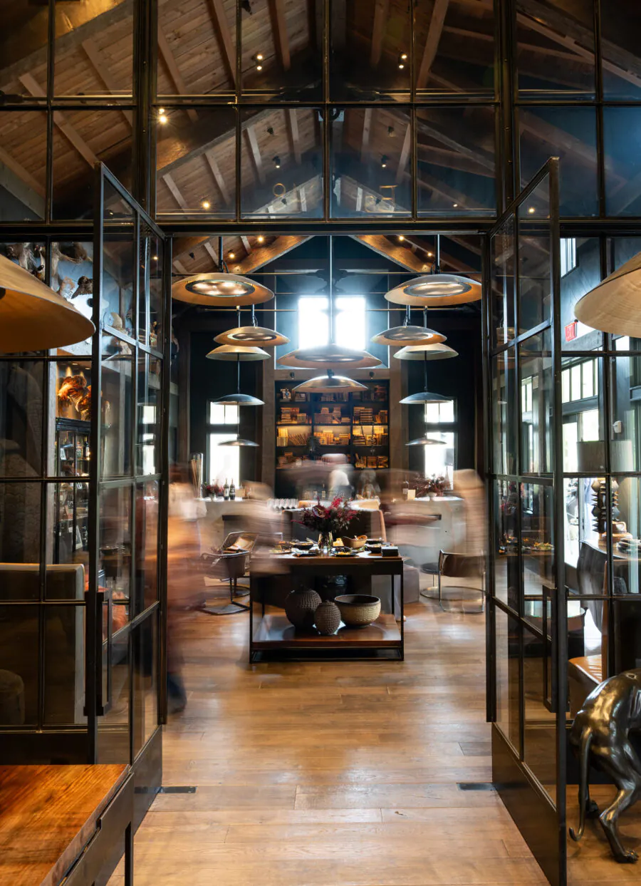 A spacious, modern interior with high wooden ceilings, glass doors, shelves of books, tables, and blurred figures walking through the room.