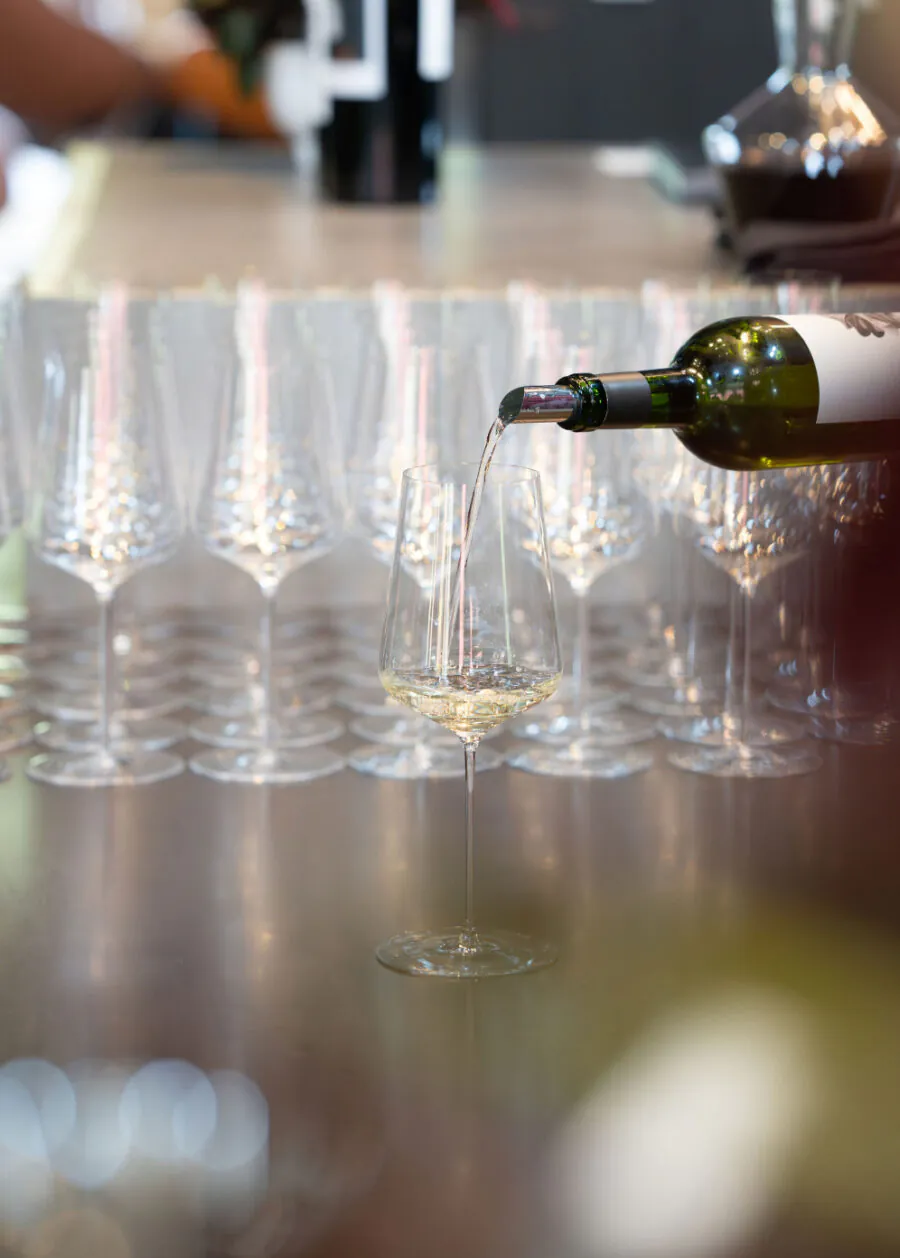 A bottle of white wine being poured into a glass, with several empty wine glasses arranged in rows on a reflective surface in the background.