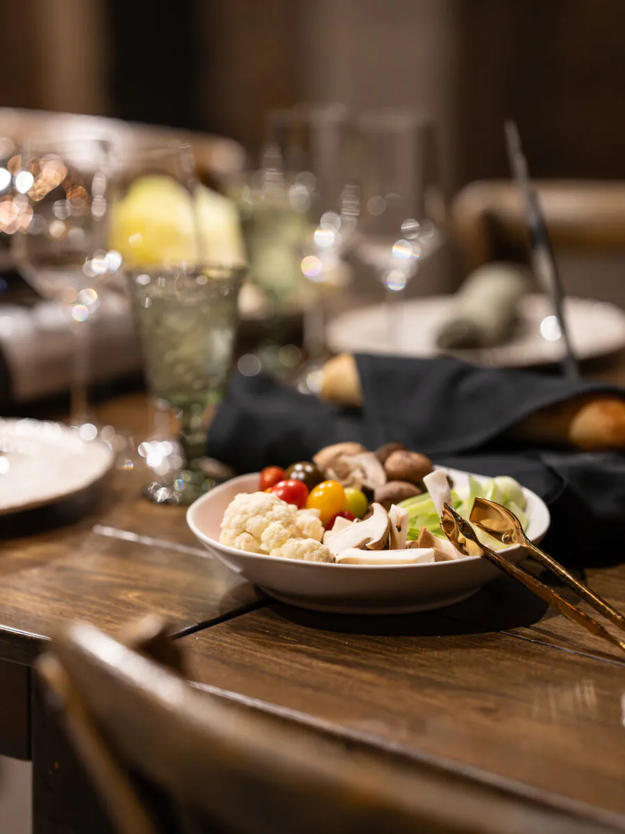 A plate of raw vegetables, including cherry tomatoes, cauliflower, mushrooms, and avocado, sits on a wooden table set with cutlery, glasses, and napkins.