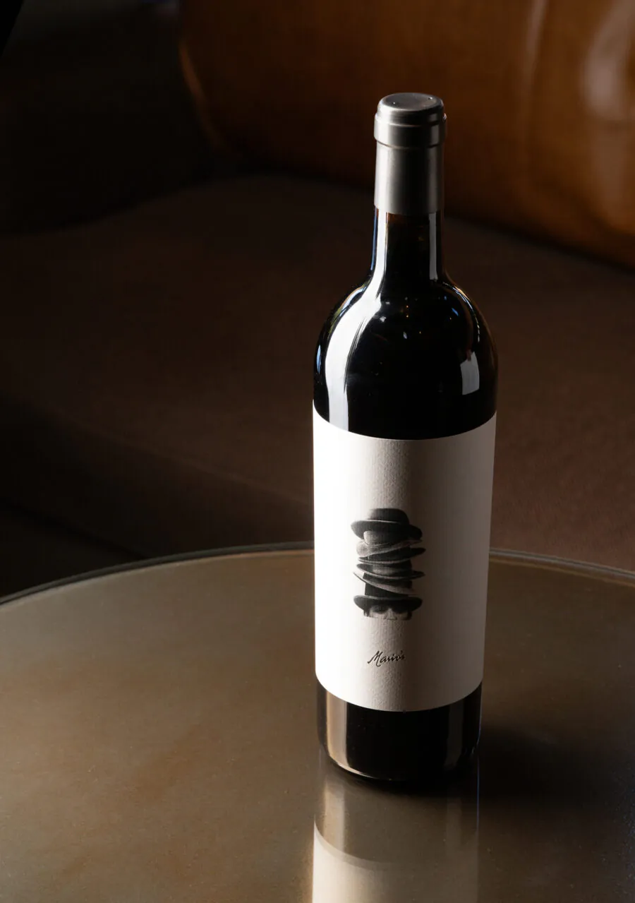 A bottle of red wine with a minimalist label stands on a round glass table, with a brown couch in the background.