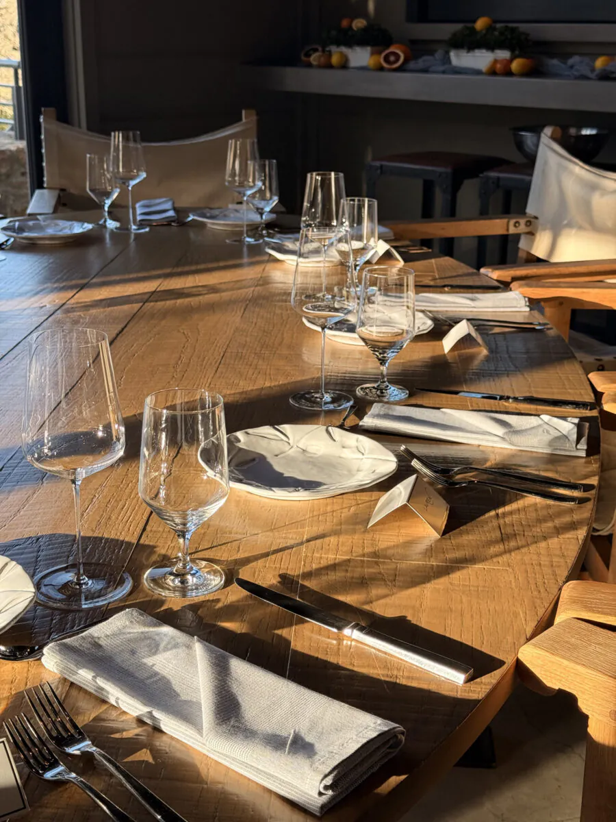 A round wooden table is set with glassware, white plates, napkins, and silverware in natural sunlight. Empty chairs surround the table.