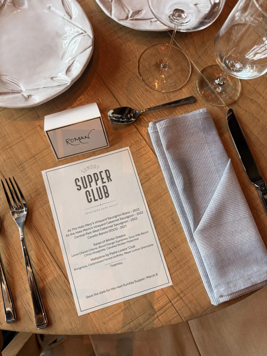 Table set for a meal with a Sunday Supper Club menu, cutlery, a napkin, wine glasses, and a small box labeled 