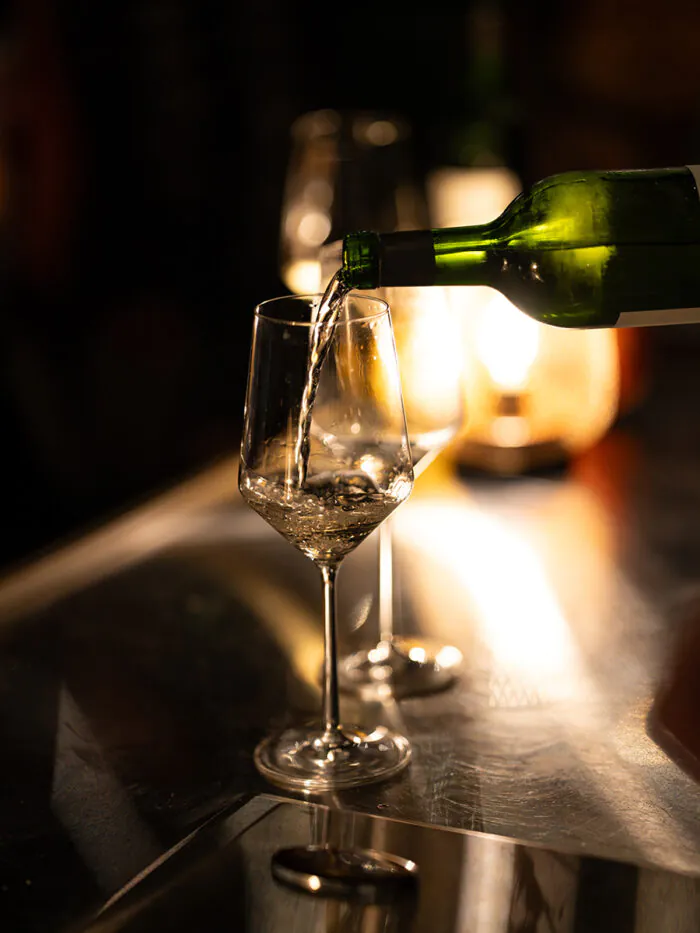 A green wine bottle pours white wine into a clear wine glass on a reflective table, with another glass and soft lighting in the background.